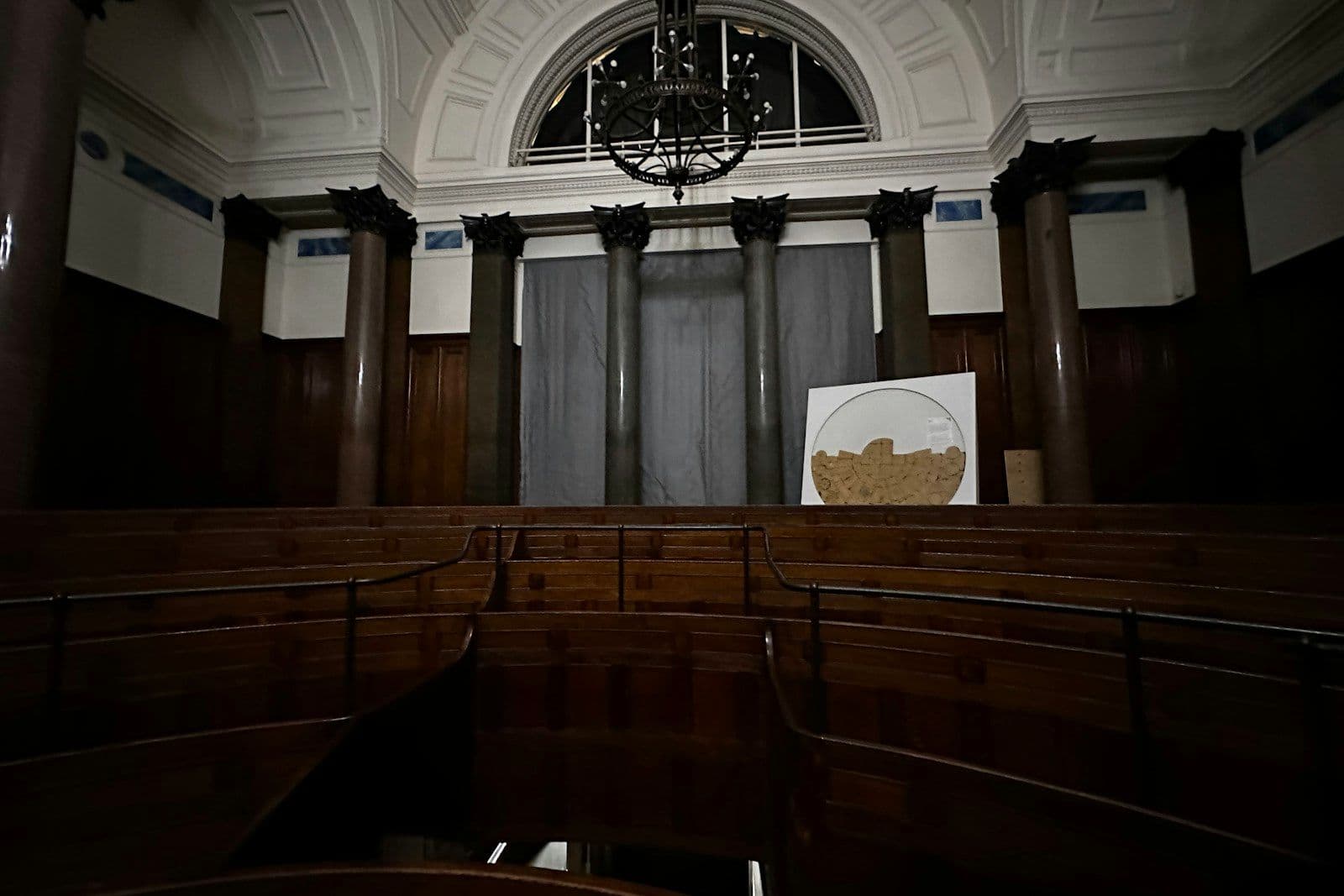 Empty wooden pews in a dim, formal hall with columns, an arched window, a hanging chandelier, and a large circular emblem on the wall