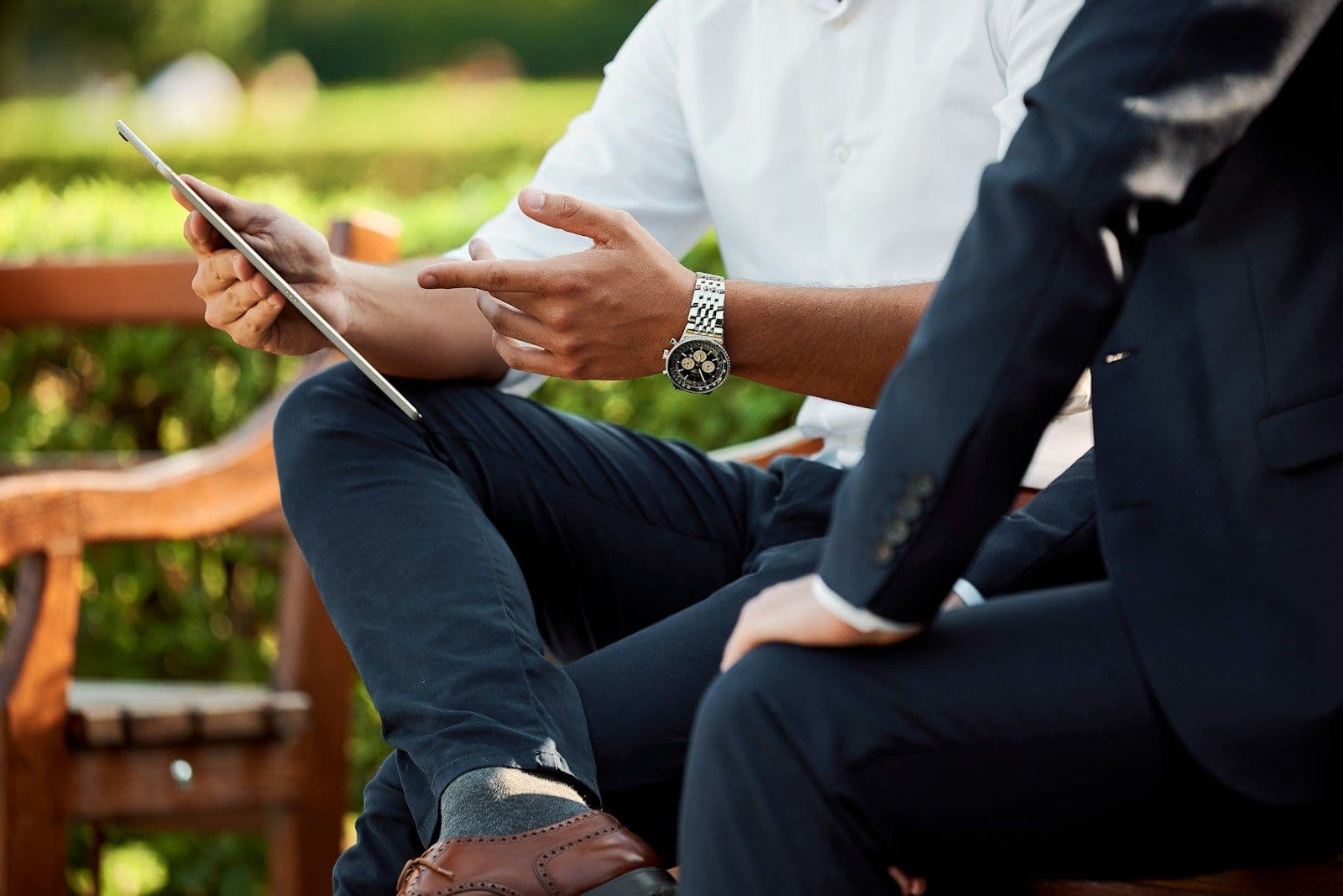 Two people seated outdoors on a bench in business attire, one holding a tablet while they talk.