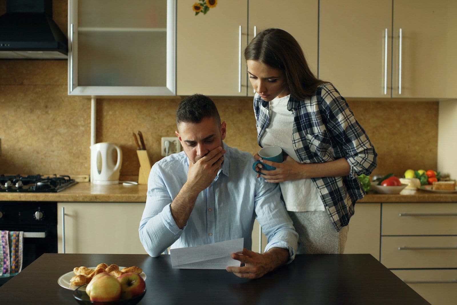 A man at a kitchen table looks worried while reading a paper as a woman stands beside him with a concerned expression.