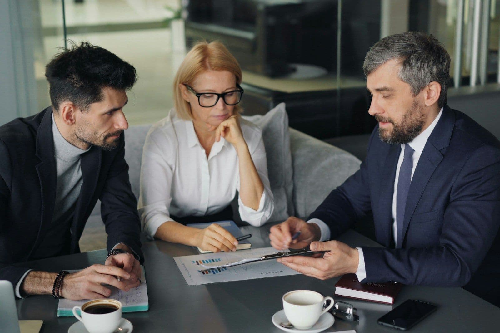 Three professionals in business attire meet at a table with coffee cups, a laptop, and a document, a man in a navy suit pointing at papers while a woman with glasses listens.