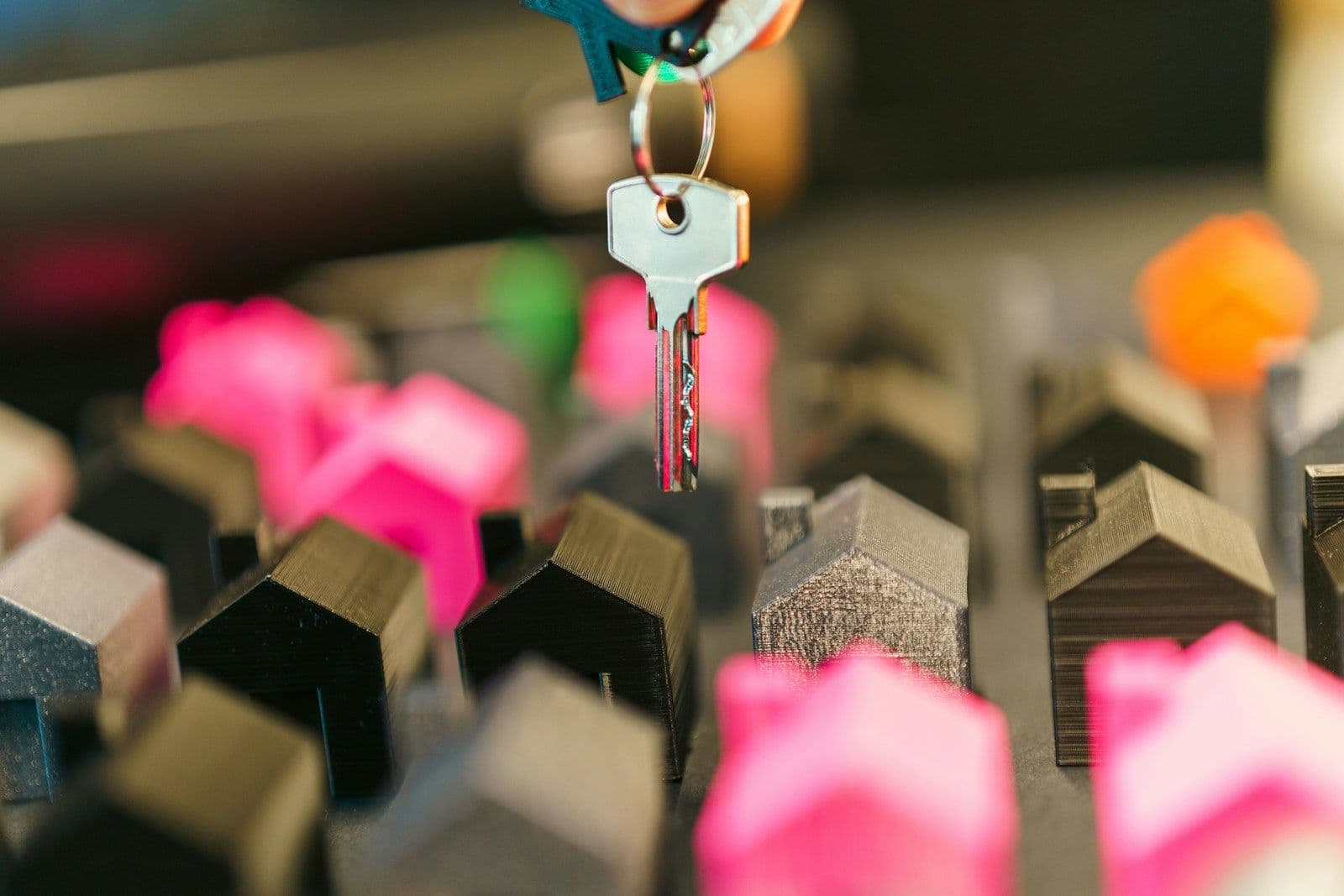 A silver house key on a colorful keyring held over a cluster of small toy houses in black, gray, and pink.
