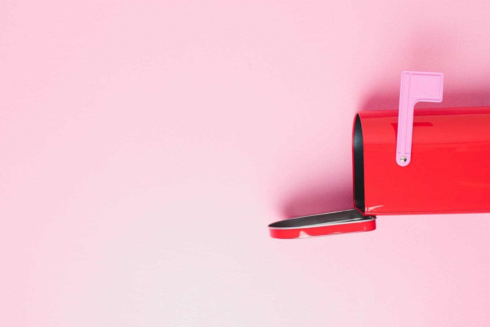 Red mailbox with open lid and small flag raised, set against a plain pink background