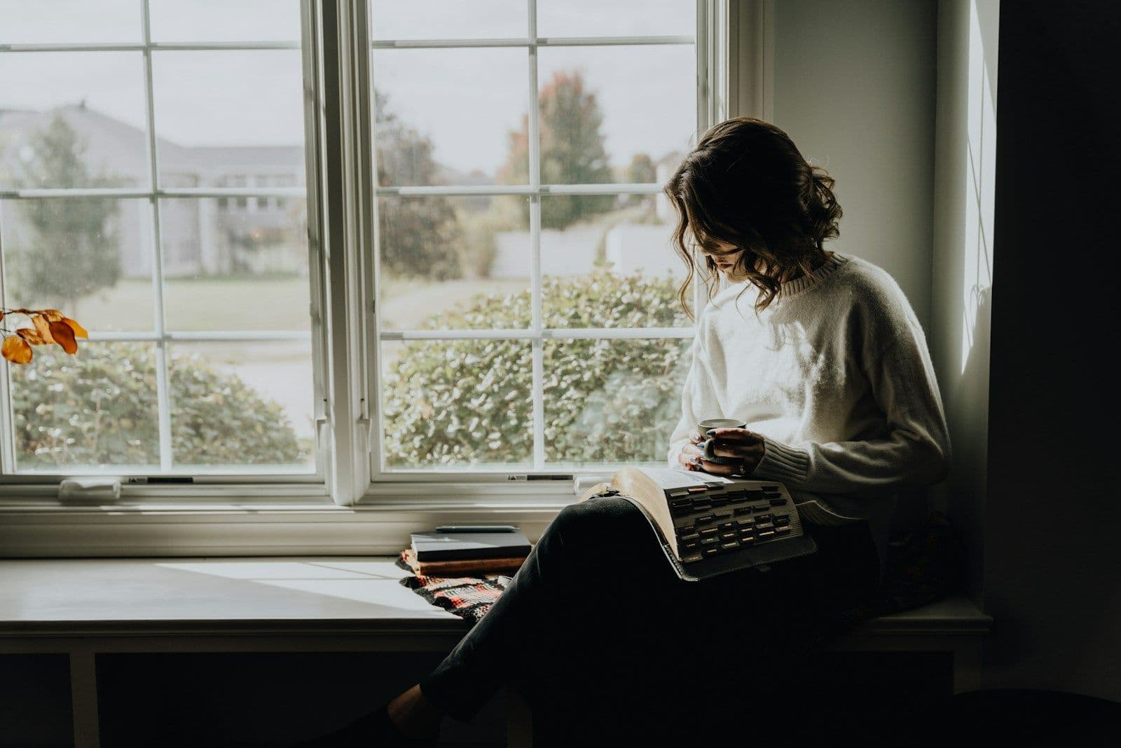 A person sits by a bright window in a white sweater, reading printed papers with a cup nearby, in a calm, quiet home setting.