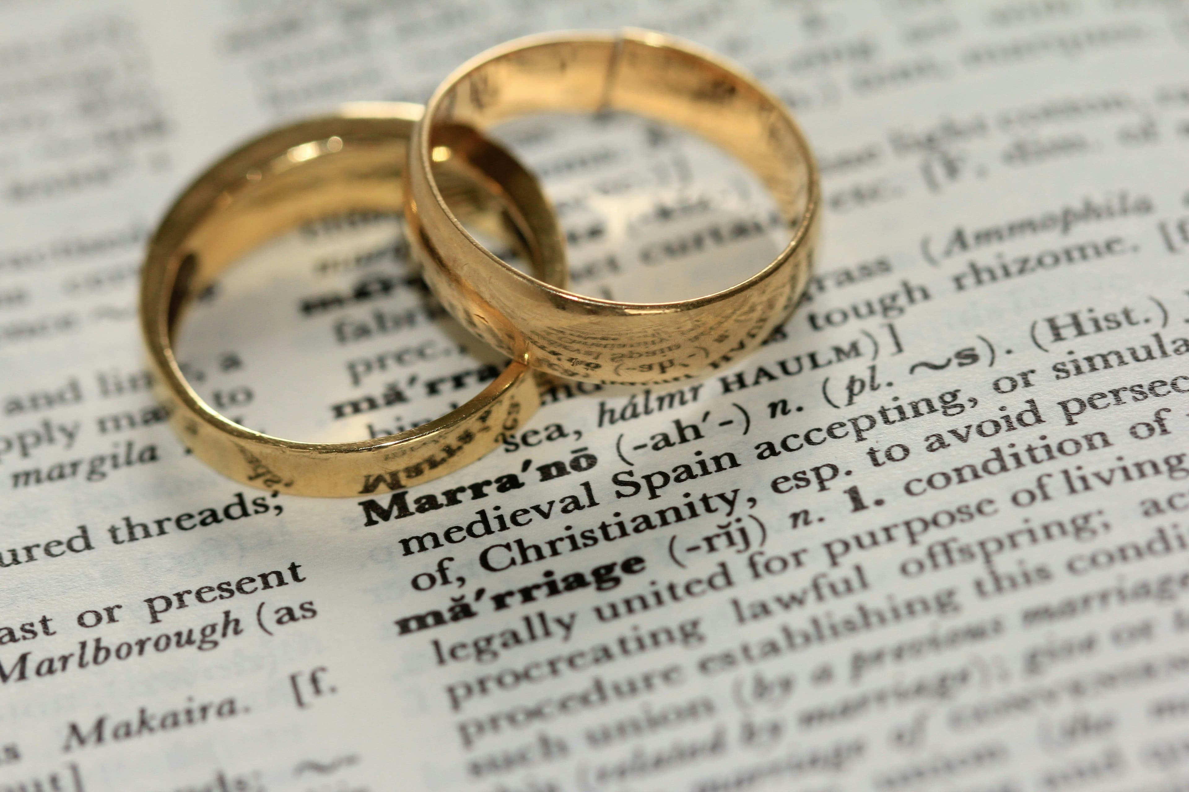 Two gold wedding rings resting on an open dictionary page with the word "marriage" visible.