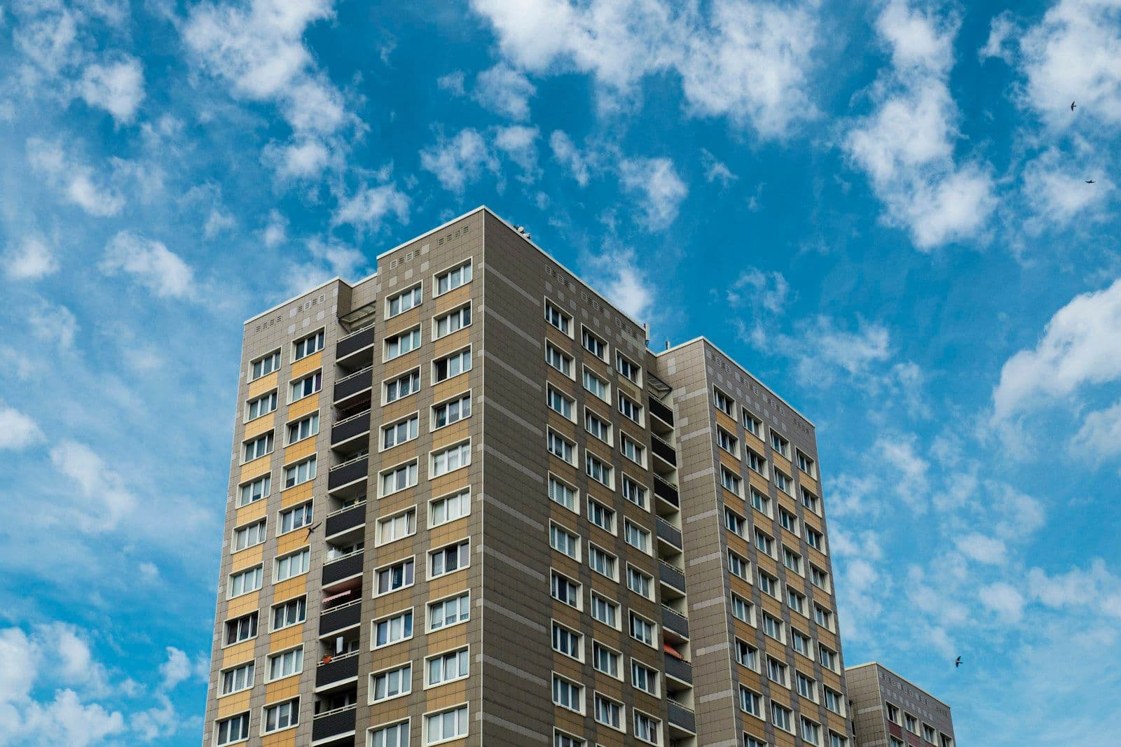 Tall beige high-rise apartment building with many windows and balconies under a bright blue sky with white clouds