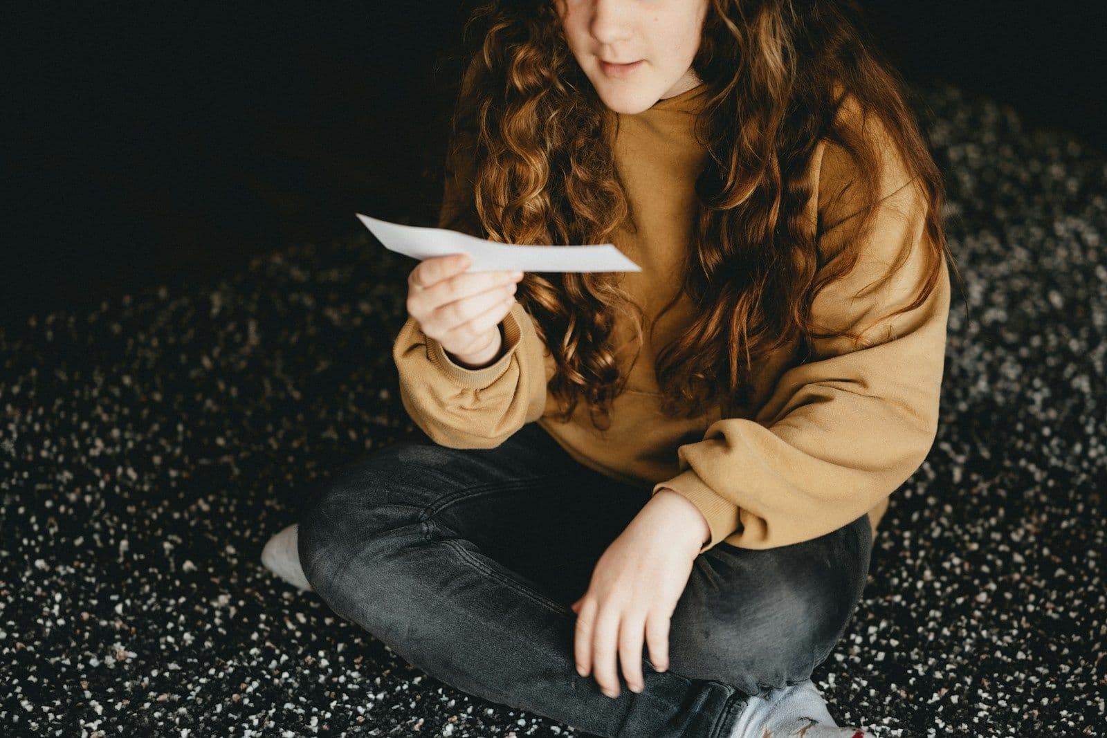 Young woman with long curly auburn hair in a mustard sweater and dark jeans sits on a speckled floor, reading a letter in her hands