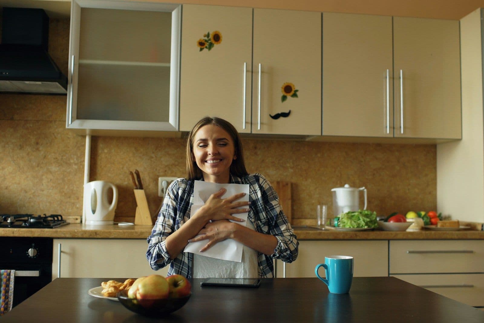 A smiling woman in a plaid shirt holds a closed white folder to her chest while standing in a bright, tidy kitchen.