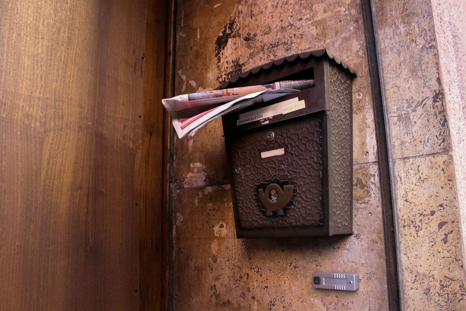 Rusty ornate metal mailbox mounted on a textured stone wall, with letters and envelopes sticking out of the slot