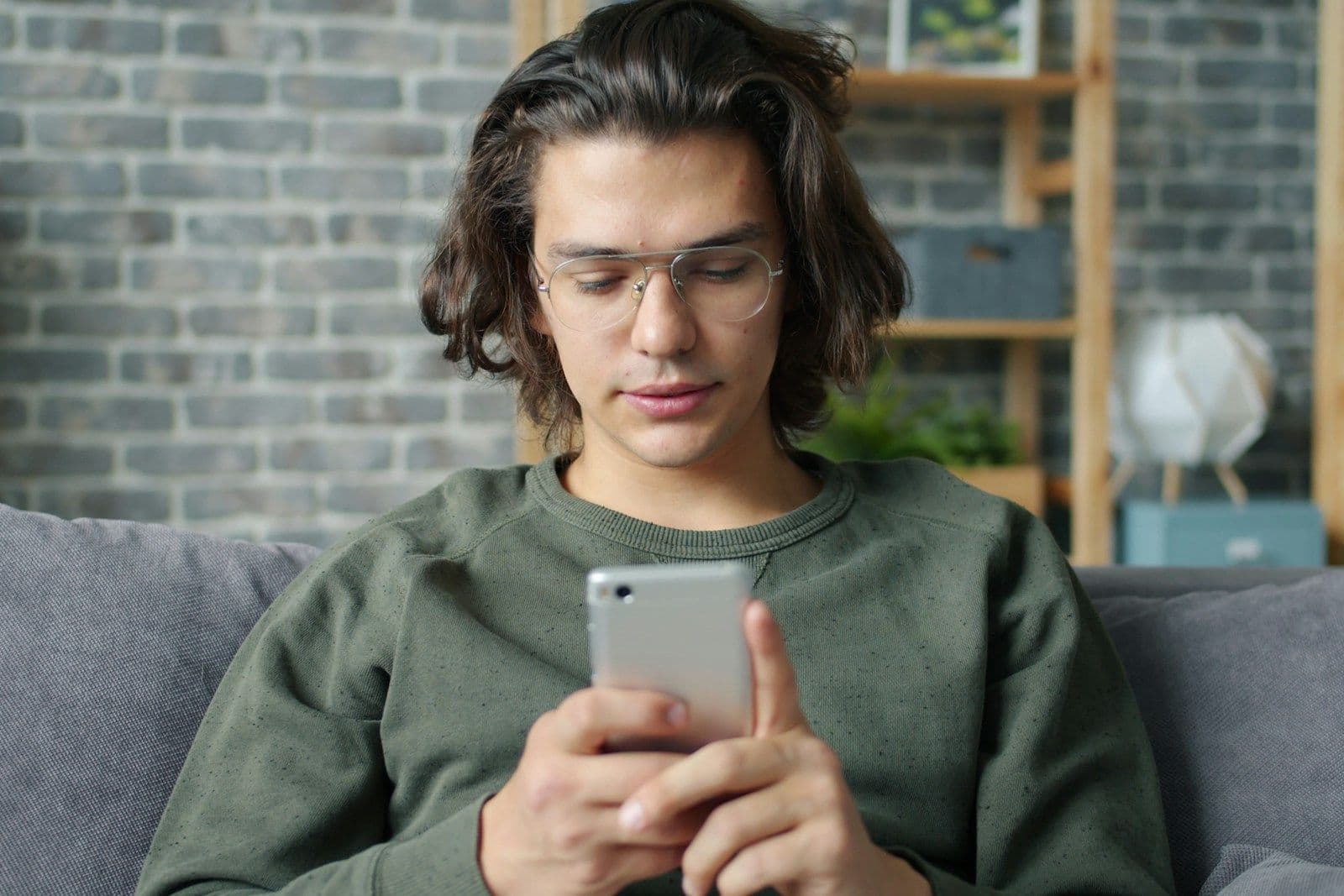 A young man with wavy dark hair and clear-framed glasses in an olive sweatshirt sits on a gray couch, looking down at a smartphone in his hands, with a brick wall and shelves behind him.