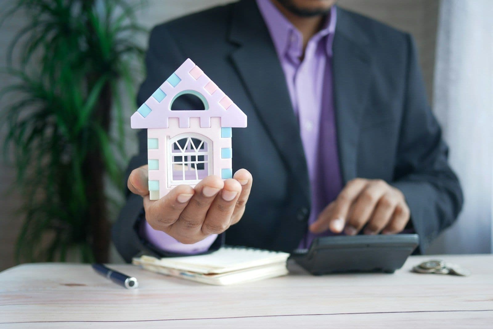 A man in a dark suit holds a small pastel pink toy house in his palm while using a calculator at a desk with a notebook and pen.