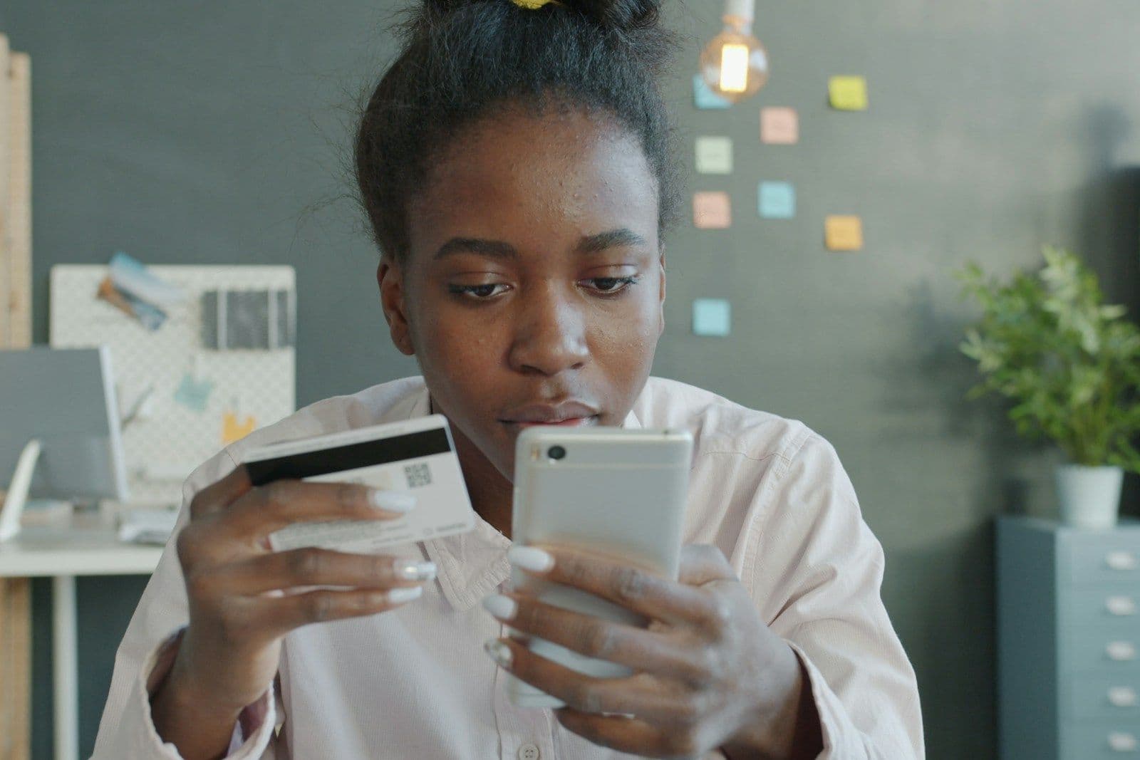 A woman in a light pink blouse holds a credit card in one hand and a smartphone in the other, looking intently at the phone in a bright office.