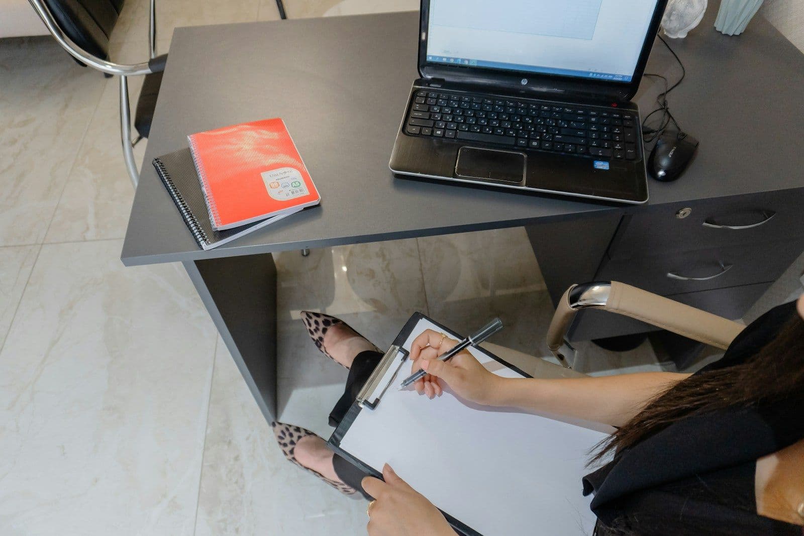 Person seated at a desk writing on a clipboard near a laptop, viewed from above
