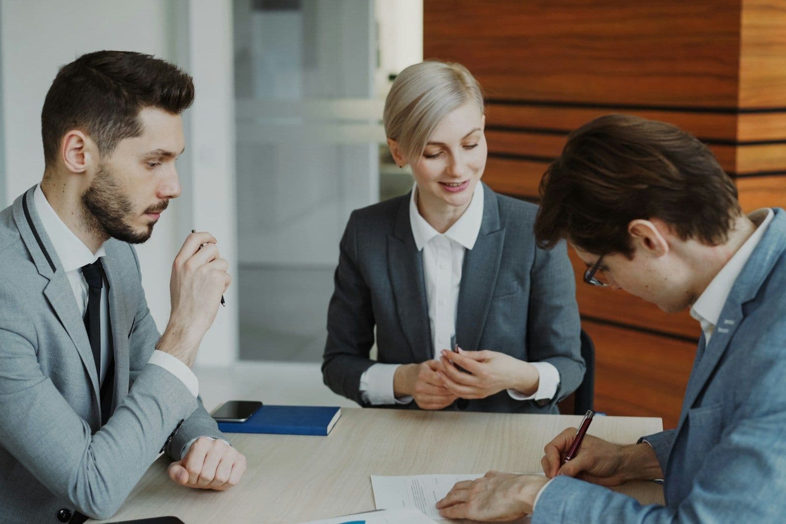 Three professionals in business attire sit at a wooden table reviewing documents, one man resting his chin on his hand while another signs papers.